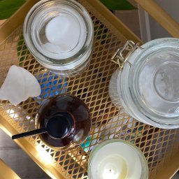 Ciałodusza - Zestaw do aromaterapii na złotej tacy: szklane słoiki z zawartością, brązowa butelka z pompką, biały kamień gua sha i zapalona świeca w szklanym pojemniku, widok z góry.