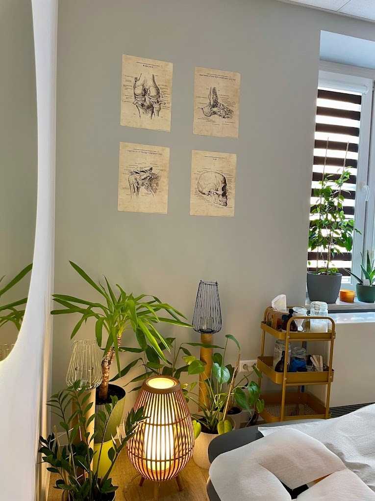Gabinet z łóżkiem do zabiegów, dekorowany roślinami i lampami, na ścianie wiszą grafiki przedstawiające anatomię stawów i czaszkę.