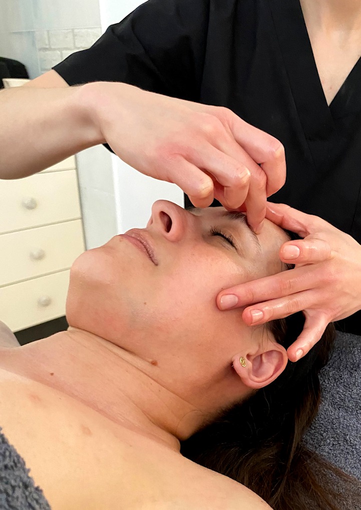 Kobieta leżąca na plecach podczas zabiegu na twarz, skupienie na masażu okolic czoła i brwi, widoczne dłonie masażysty w czarnym uniformie.