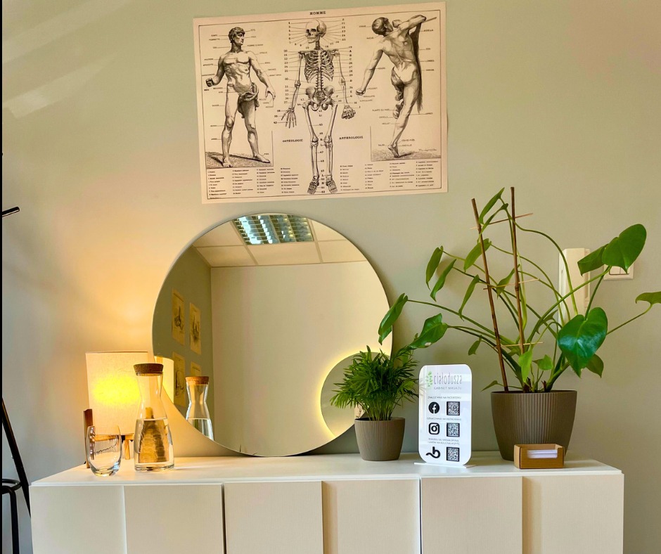Jasne wnętrze gabinetu z okrągłym lustrem z podświetleniem LED, plakatem anatomicznym na ścianie, roślinami doniczkowymi i szklanym dzbankiem z wodą, tworzące relaksującą atmosferę.