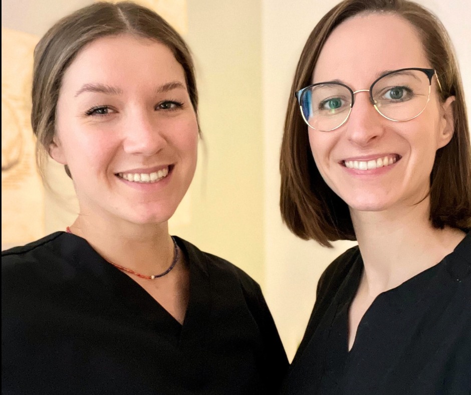 Portret dwóch uśmiechniętych kobiet w czarnych uniformach, prawdopodobnie pracownice salonu, jedna w okularach, obie z delikatnym makijażem, stojące na tle jasnej ściany.