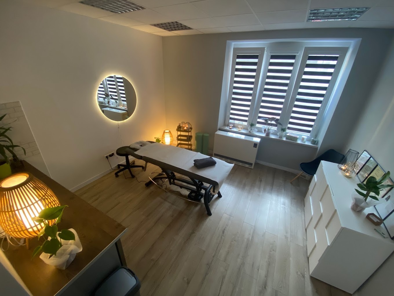 Jasne wnętrze gabinetu z łóżkiem do zabiegów, okrągłym lustrem z podświetleniem i drewnianą podłogą, widziane z góry.