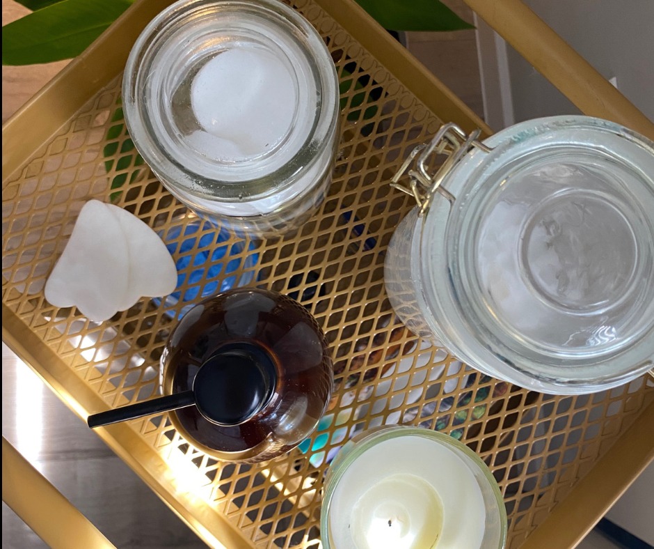 Zestaw do aromaterapii na złotej tacy: szklane słoiki z zawartością, brązowa butelka z pompką, biały kamień gua sha i zapalona świeca w szklanym pojemniku, widok z góry.