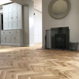 Cyklinomania.pl - Jasna podłoga z parkietu ułożonego we wzór jodełki francuskiej w salonie z szarymi ścianami, kominkiem z kratą i szarą szafą w tle.