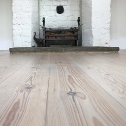 Cyklinomania.pl - Jasne, odnowione deski podłogowe z widoczną strukturą drewna, w tle biały ceglany kominek z żeliwnym koszem na drewno i figurką słonia