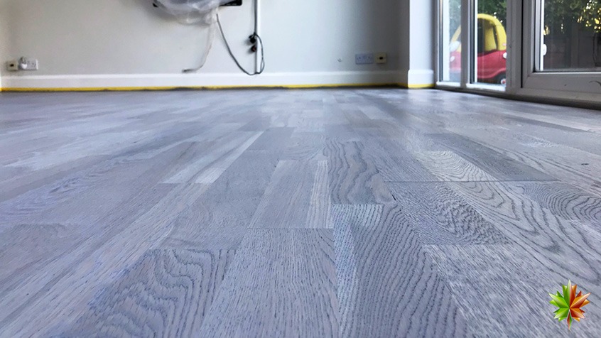 Jasnoszara, odnowiona podłoga drewniana w trakcie remontu, zabezpieczona taśmą malarską przy ścianach, z widocznymi gniazdkami elektrycznymi i elementami instalacji zakrytymi folią.