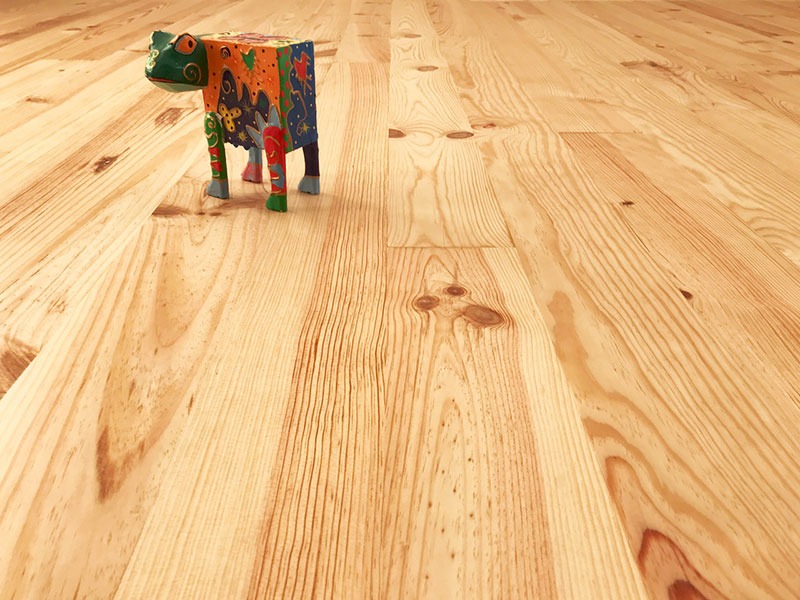 Jasna, drewniana podłoga z widocznymi słojami i małymi sękami, na której stoi kolorowa, drewniana figurka żaby.