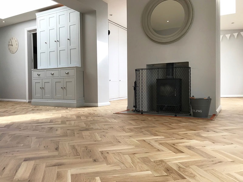 Jasna podłoga z parkietu ułożonego we wzór jodełki francuskiej w salonie z szarymi ścianami, kominkiem z kratą i szarą szafą w tle.