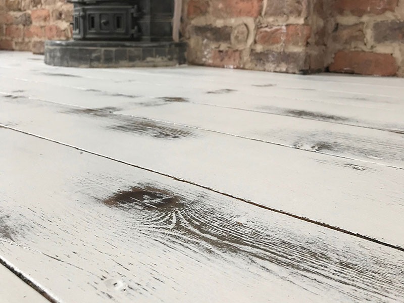 Biała, postarzana podłoga z desek w zbliżeniu, z widocznym piecem kaflowym i fragmentem ceglanej ściany w tle.