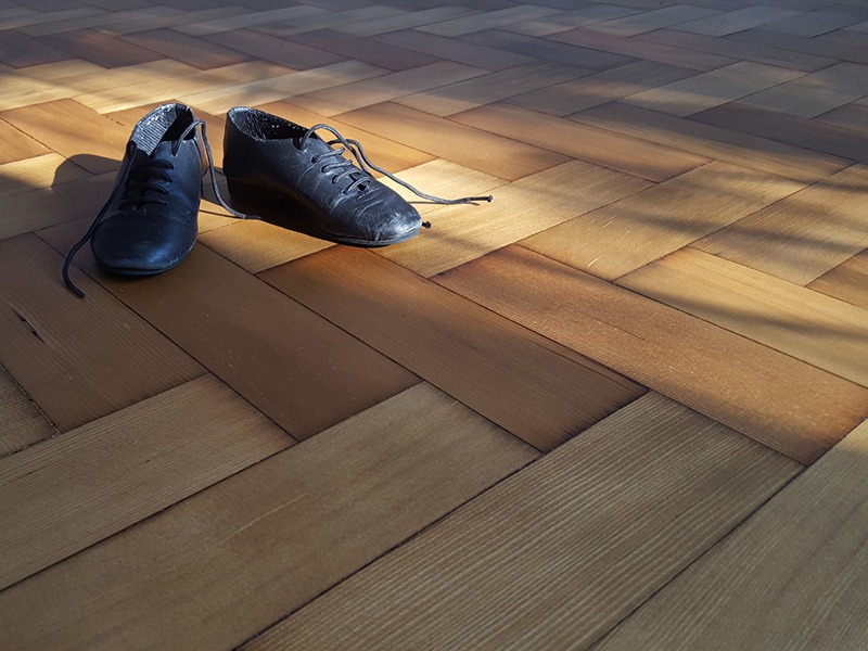 Parkiet ułożony w jodełkę, oświetlony promieniami słońca, z parą czarnych, sznurowanych butów tanecznych na wierzchu.