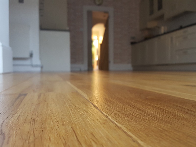 Błyszcząca, świeżo położona podłoga z paneli drewnianych w perspektywie od podłogi, z widocznym przejściem do kuchni w tle.