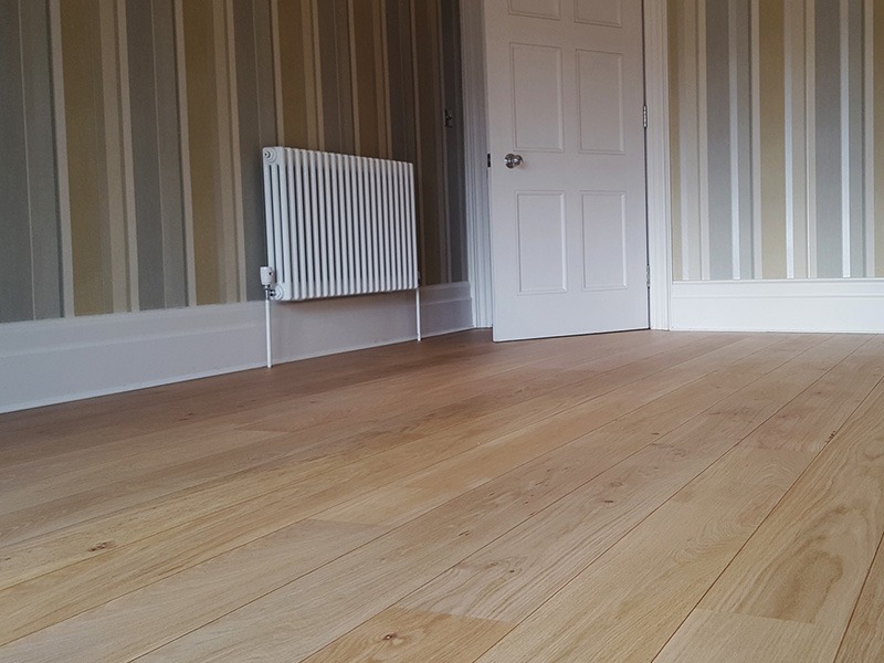 Jasna, drewniana podłoga z widocznymi słojami, ułożona w pokoju z grzejnikiem i tapetą w pionowe pasy, częściowo otwarte białe drzwi w tle.