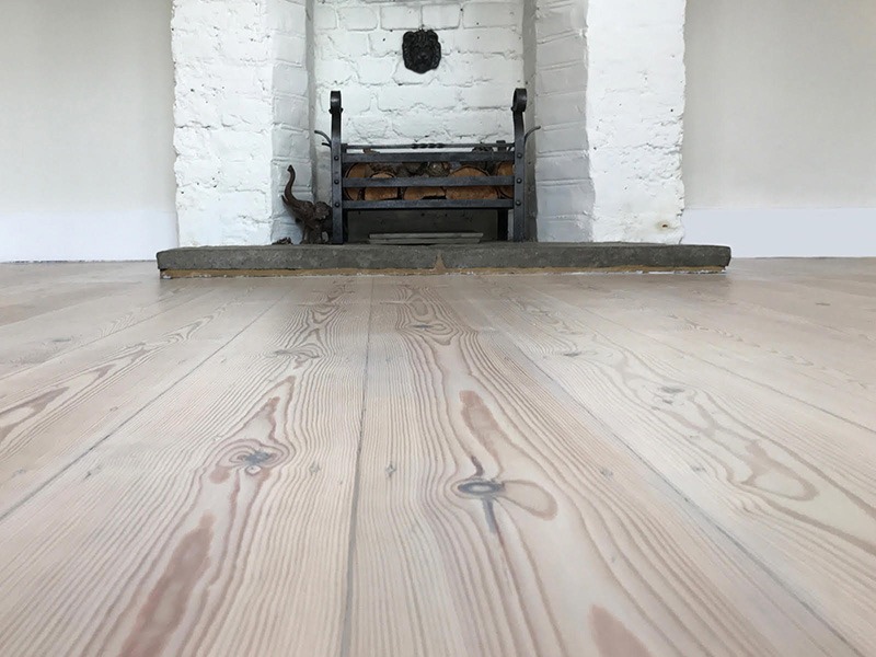 Jasne, odnowione deski podłogowe z widoczną strukturą drewna, w tle biały ceglany kominek z żeliwnym koszem na drewno i figurką słonia