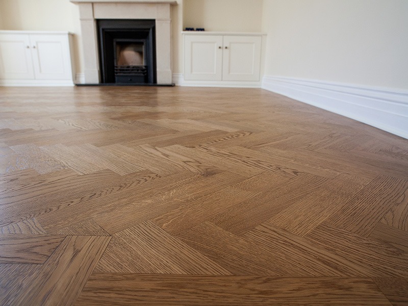 Ułożona podłoga z parkietu w jodełkę, lakierowana na brązowo, widok na kominek i białe szafki w tle.