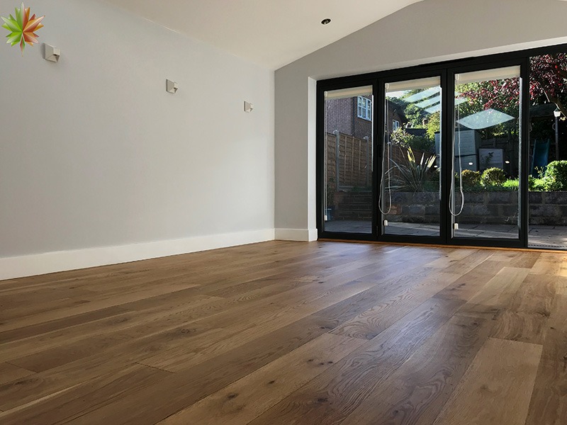 Jasny pokój z nową, brązową podłogą z desek, widok na ogród przez duże, składane, czarne drzwi balkonowe.