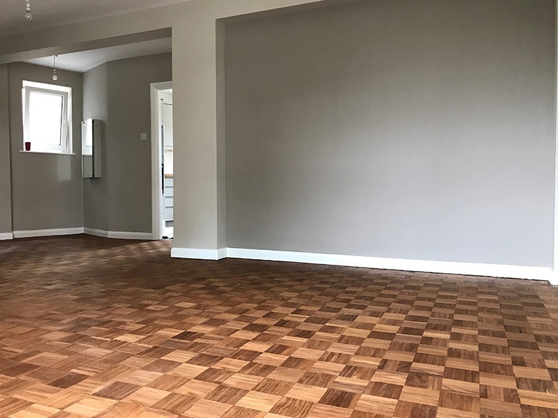 Wnętrze pokoju z parkietem ułożonym we wzór szachownicy, szare ściany i białe listwy przypodłogowe, widok na fragment kuchni w tle.