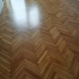 Parkiet ułożony w jodełkę po renowacji, widoczne odbicie światła z okna. Jasne drewno, świeżo polakierowane, brak listew przypodłogowych.