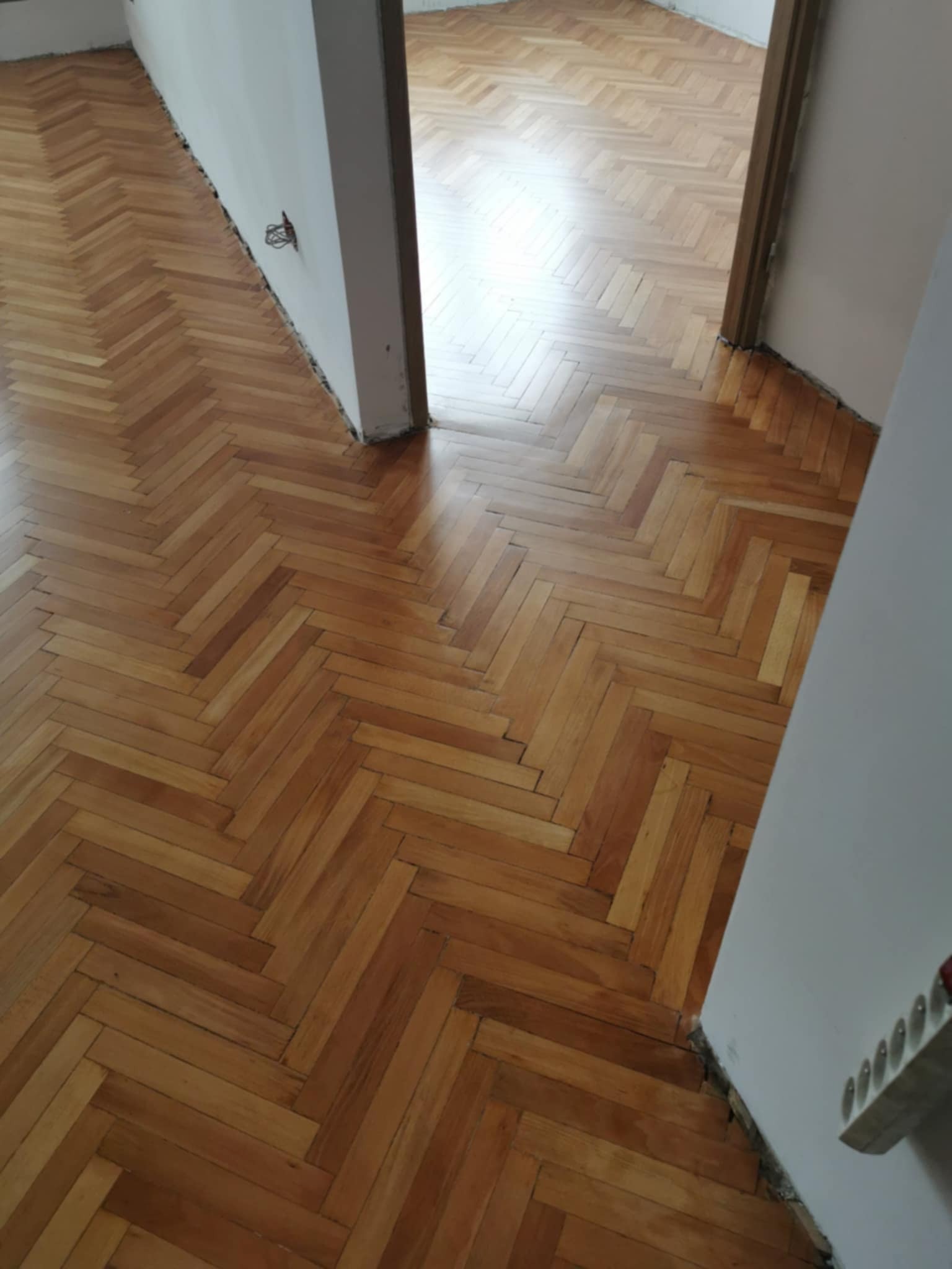 Parkiet ułożony w jodełkę, po renowacji. Widoczne przejście do innego pomieszczenia. Świeżo wycyklinowana podłoga z drewna, jasne odcienie brązu i beżu.