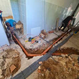 Zakład Instalacji sanitarnych, c.o. i gazu - Instalacja kanalizacyjna w trakcie montażu: widoczne pomarańczowe rury kanalizacyjne prowadzone w wykopie w podłodze, obok wiązka szarych rur osłonowych, na ścianie widoczne płytki ceramiczne,...