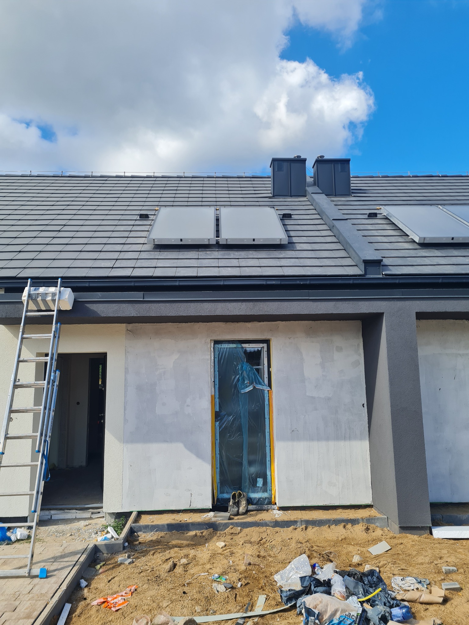 Nowy dom w trakcie wykończenia, widoczne panele słoneczne na dachu, drabina oparta o ścianę, okno zabezpieczone folią, teren wokół domu w trakcie prac ziemnych.