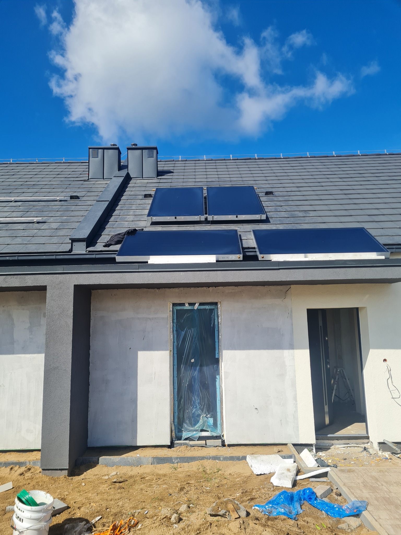 Nowoczesny dom jednorodzinny z panelami słonecznymi na grafitowym dachu, widoczne kominy i okna zabezpieczone folią, teren wokół budynku w trakcie prac wykończeniowych.