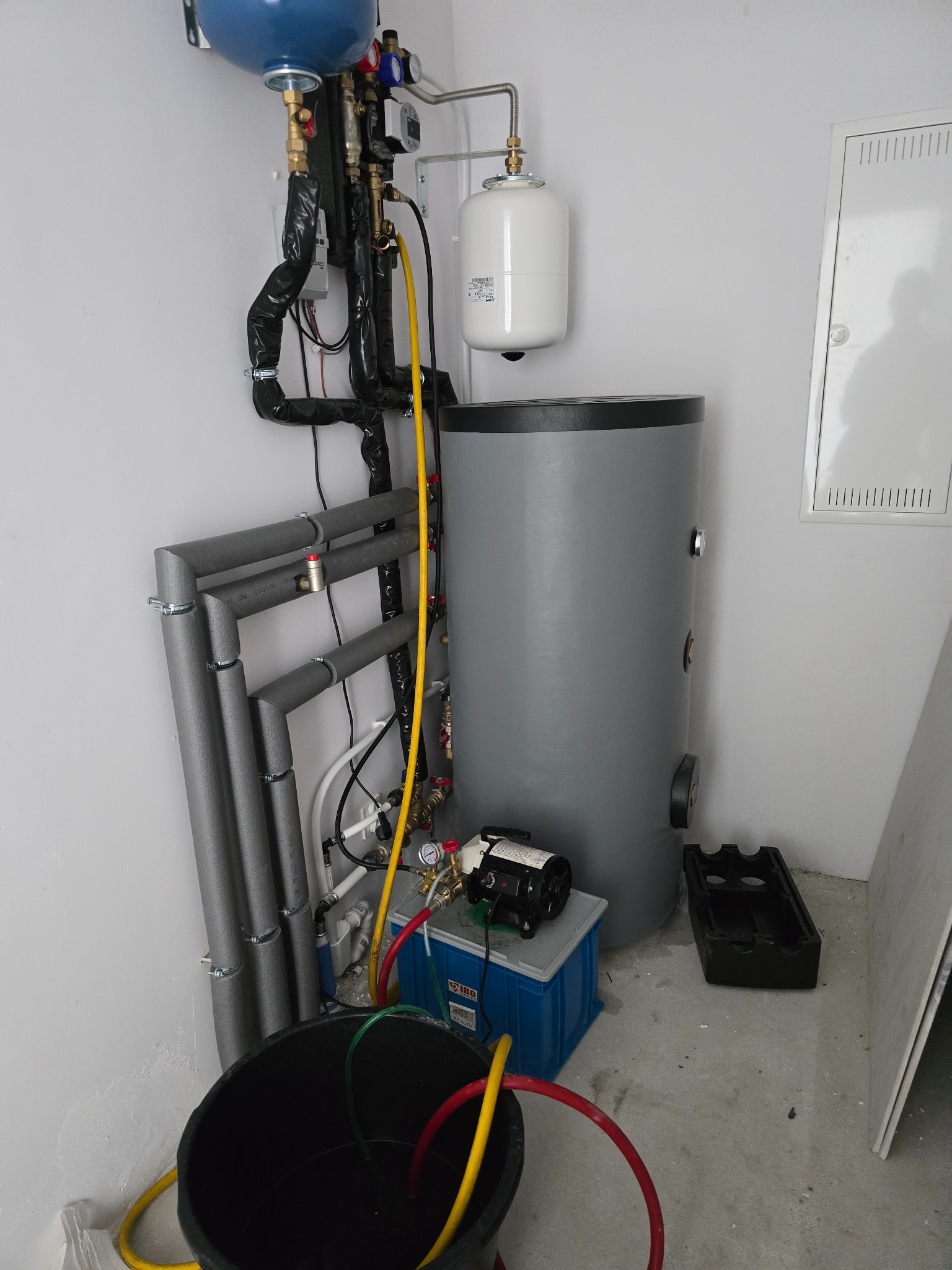 Szary bojler z podłączeniami hydraulicznymi i elektrycznymi w kotłowni, widoczna pompa, rury z izolacją, zbiornik wyrównawczy, oraz wiadro z wężami.