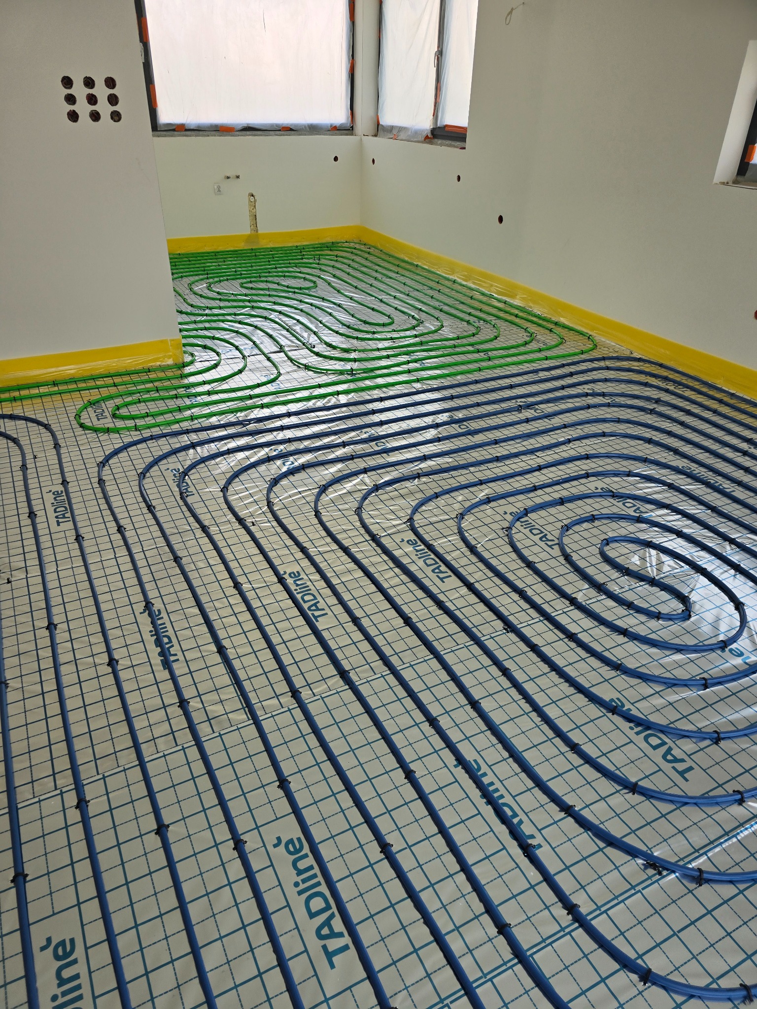 Niebieskie i zielone rury ogrzewania podłogowego marki Tadline ułożone w pętle na białej folii z siatką, żółta folia ochronna przy ścianach, surowe wnętrze w trakcie budowy.