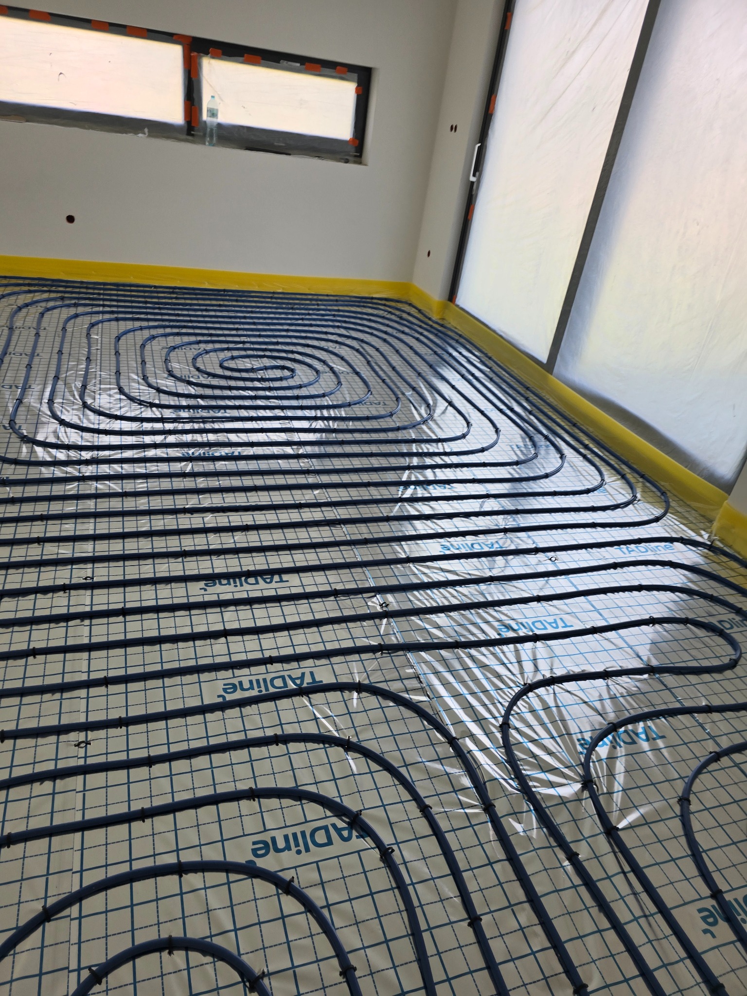 Nisko kątowe ujęcie podłogówki z niebieskich rur na macie izolacyjnej z nadrukiem TADIline, zabezpieczone żółtą folią przy ścianach, widoczny fragment okna i drzwi zabezpieczonych folią malarską.