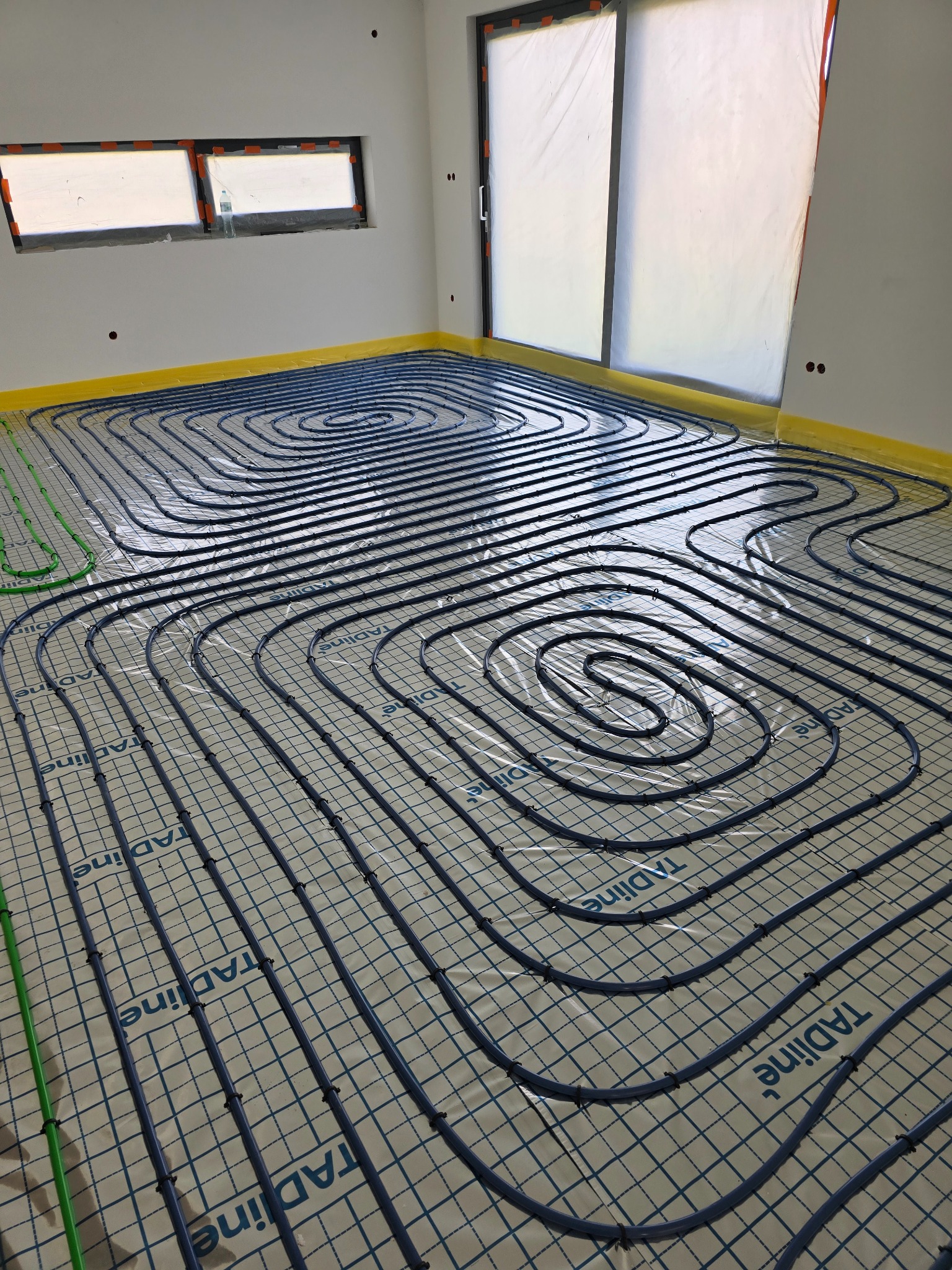 Niebieskie rury ogrzewania podłogowego ułożone spiralnie na siatce montażowej w jasnym pomieszczeniu z oknami, zabezpieczone folią i taśmą malarską przy ścianach.