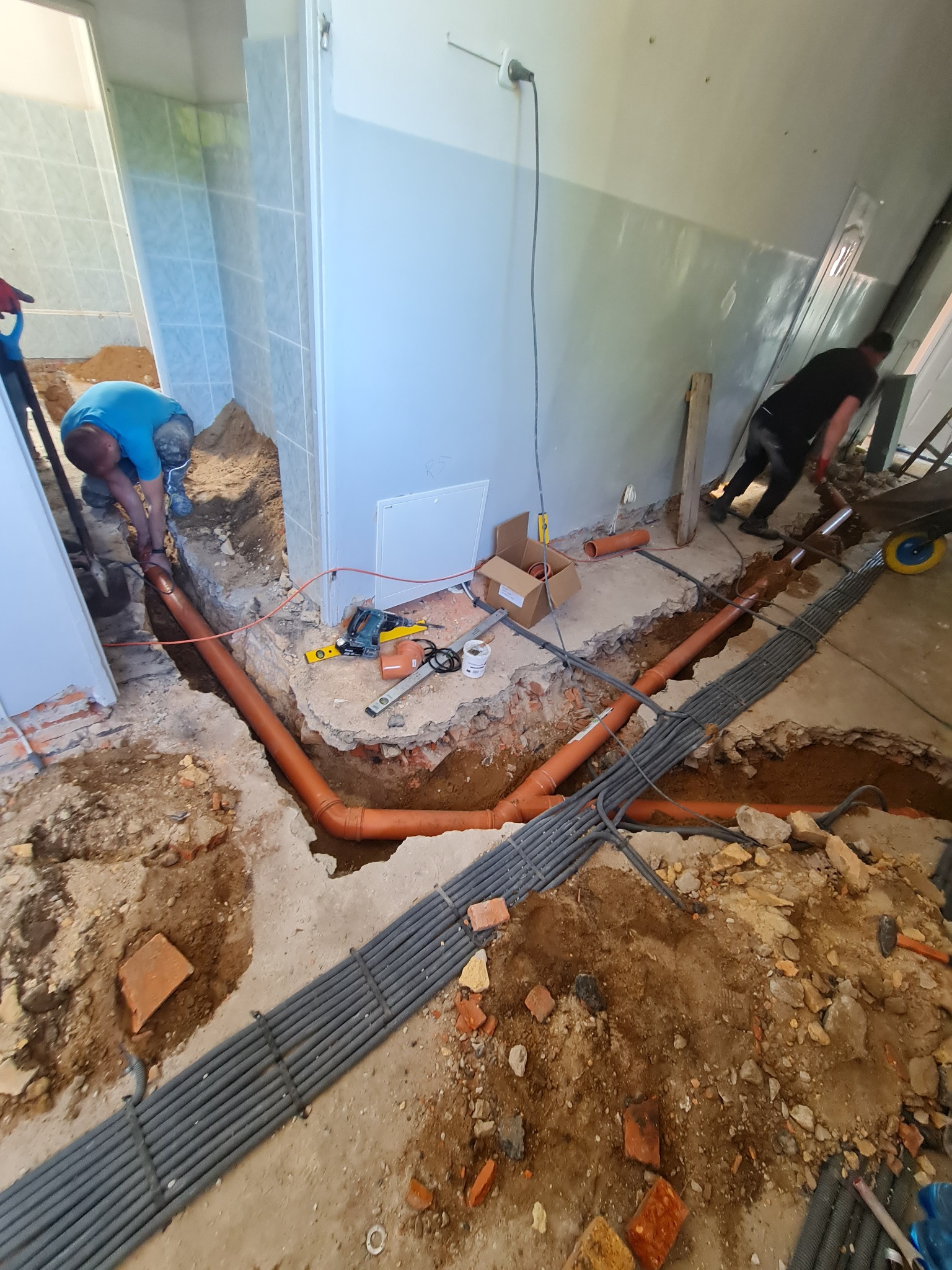 Instalacja kanalizacyjna w trakcie montażu: widoczne pomarańczowe rury kanalizacyjne prowadzone w wykopie w podłodze, obok wiązka szarych rur osłonowych, na ścianie widoczne płytki ceramiczne,...