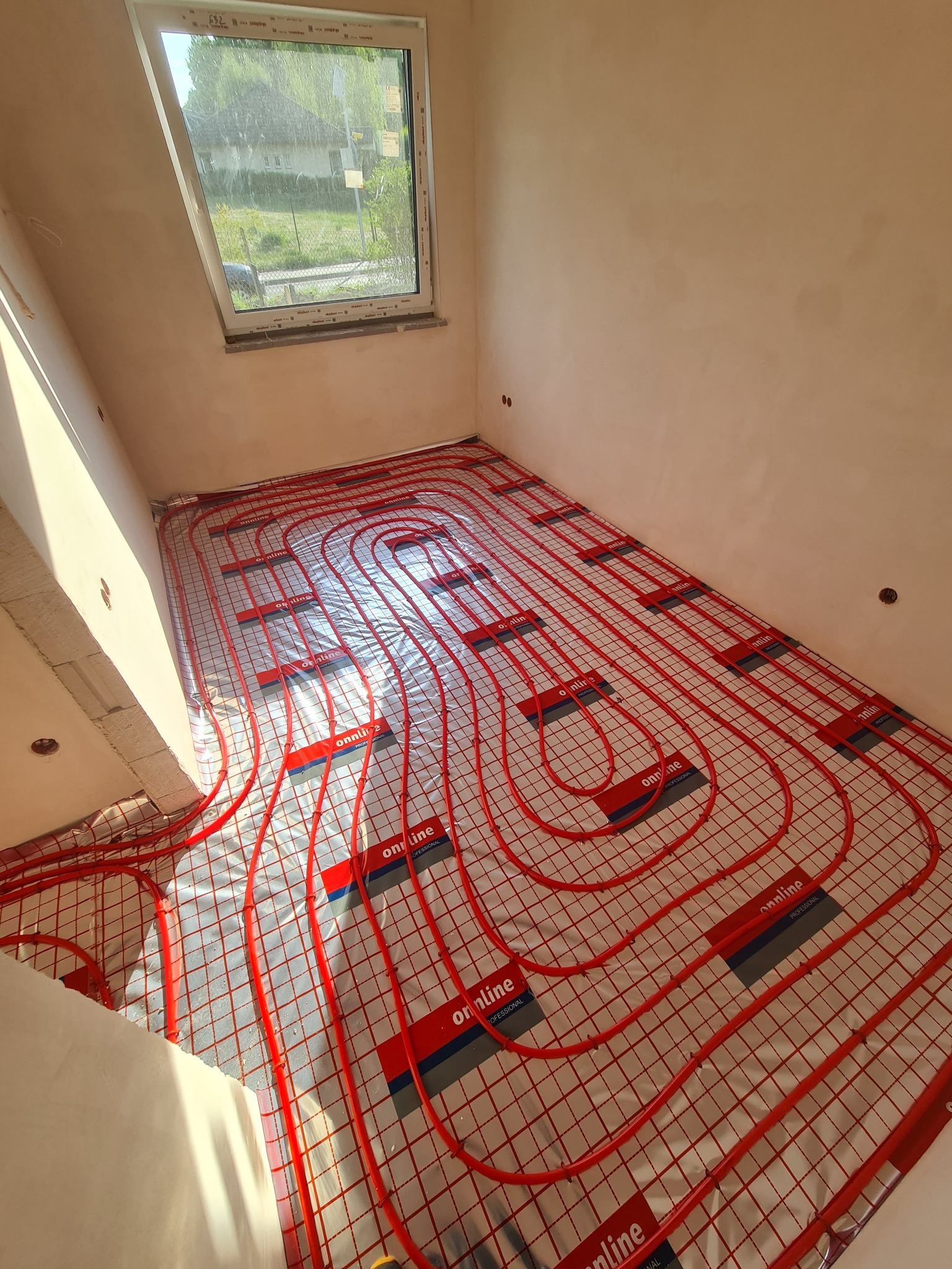 Widok z góry na rozłożoną instalację ogrzewania podłogowego z czerwonych rur na folii z nadrukiem w nowym budynku w trakcie budowy, z oknem dachowym w tle.