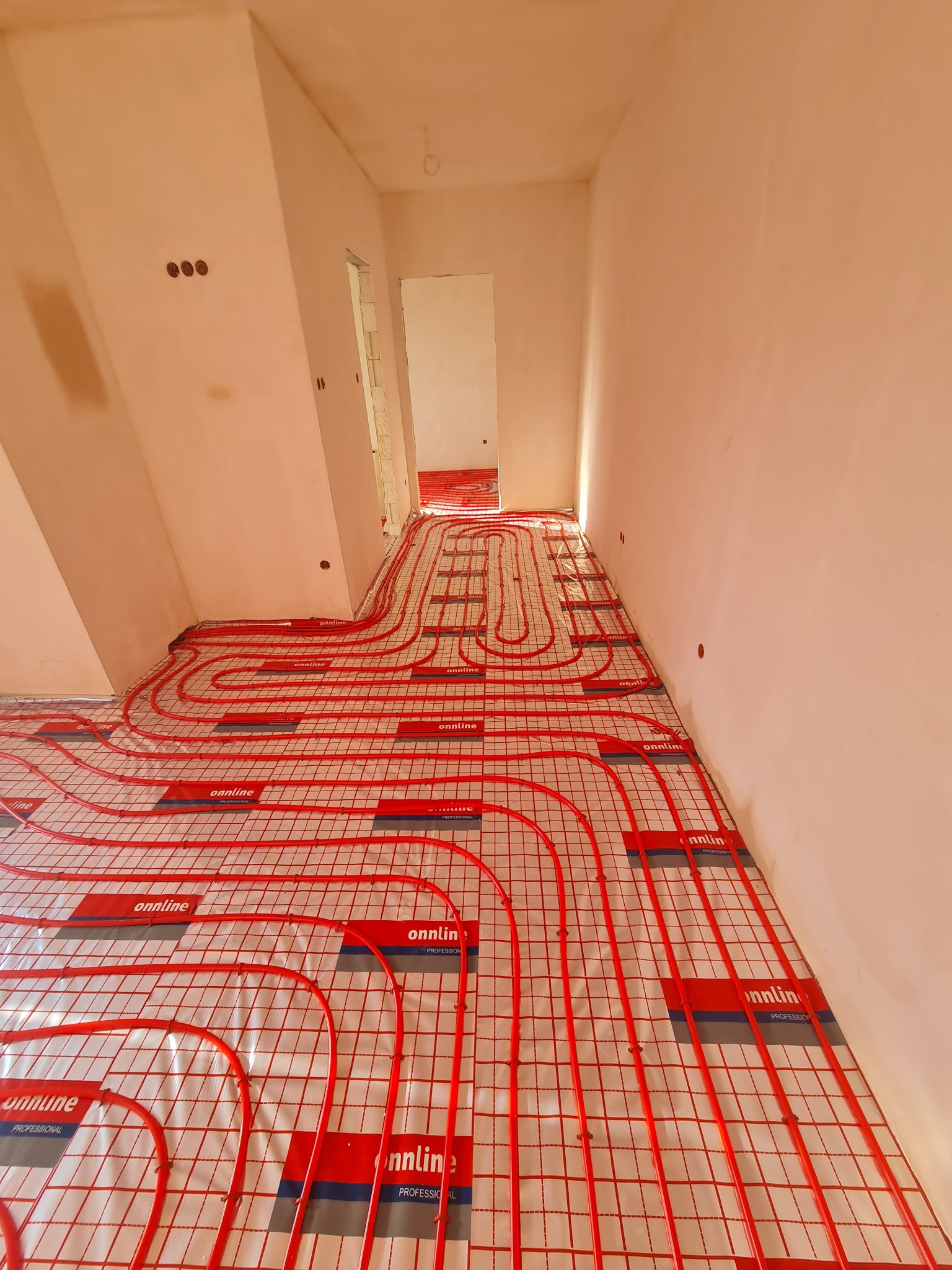 Czerwone rury ogrzewania podłogowego ułożone na białej folii z nadrukiem w surowym wnętrzu w trakcie instalacji, widoczne otwory w ścianie.