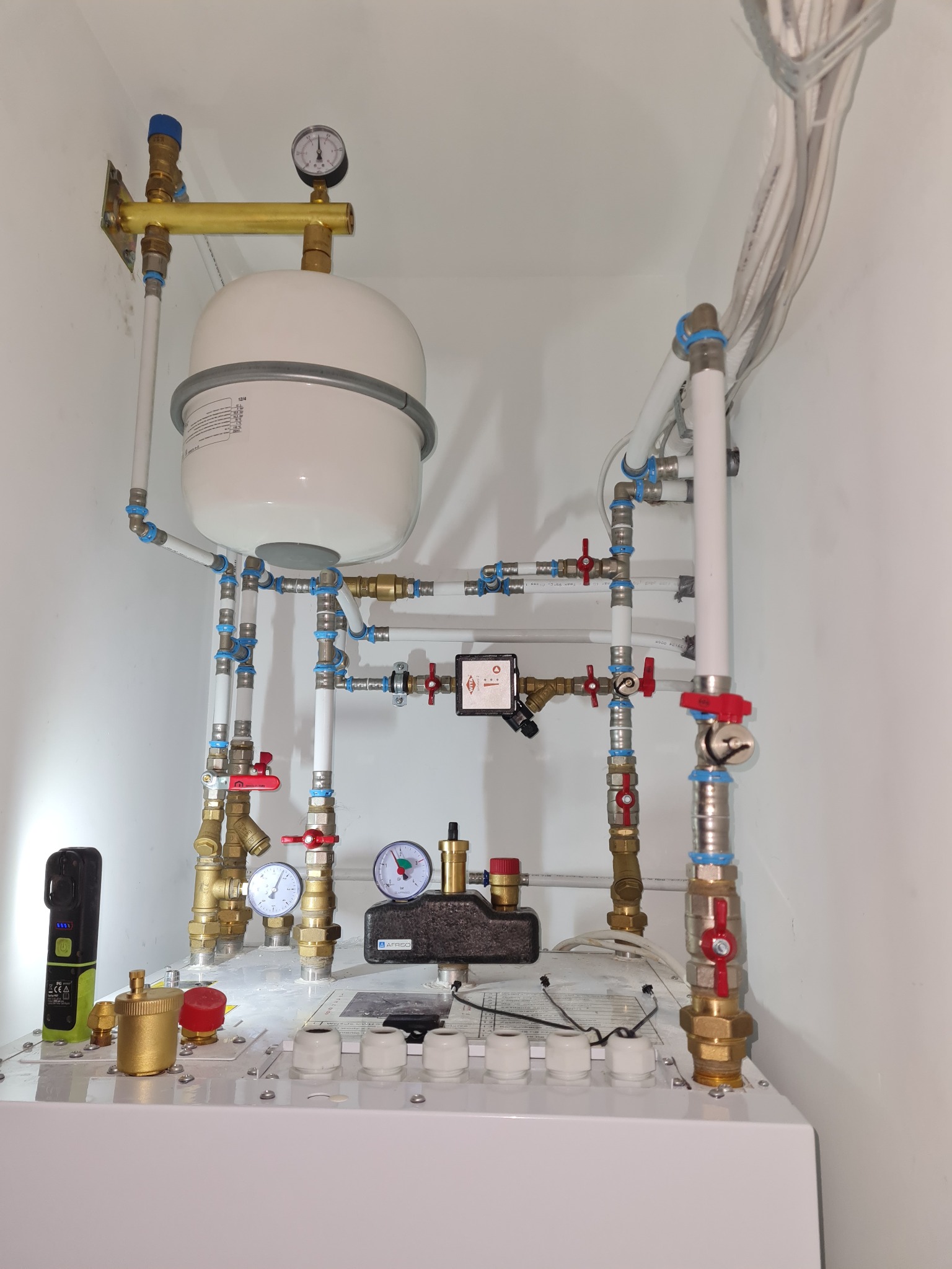 Rozbudowana instalacja hydrauliczna z białym zbiornikiem wyrównawczym, licznymi zaworami, manometrami i rurami z tworzywa sztucznego oraz mosiądzu, zamontowana na białej ścianie w pomieszczeniu...