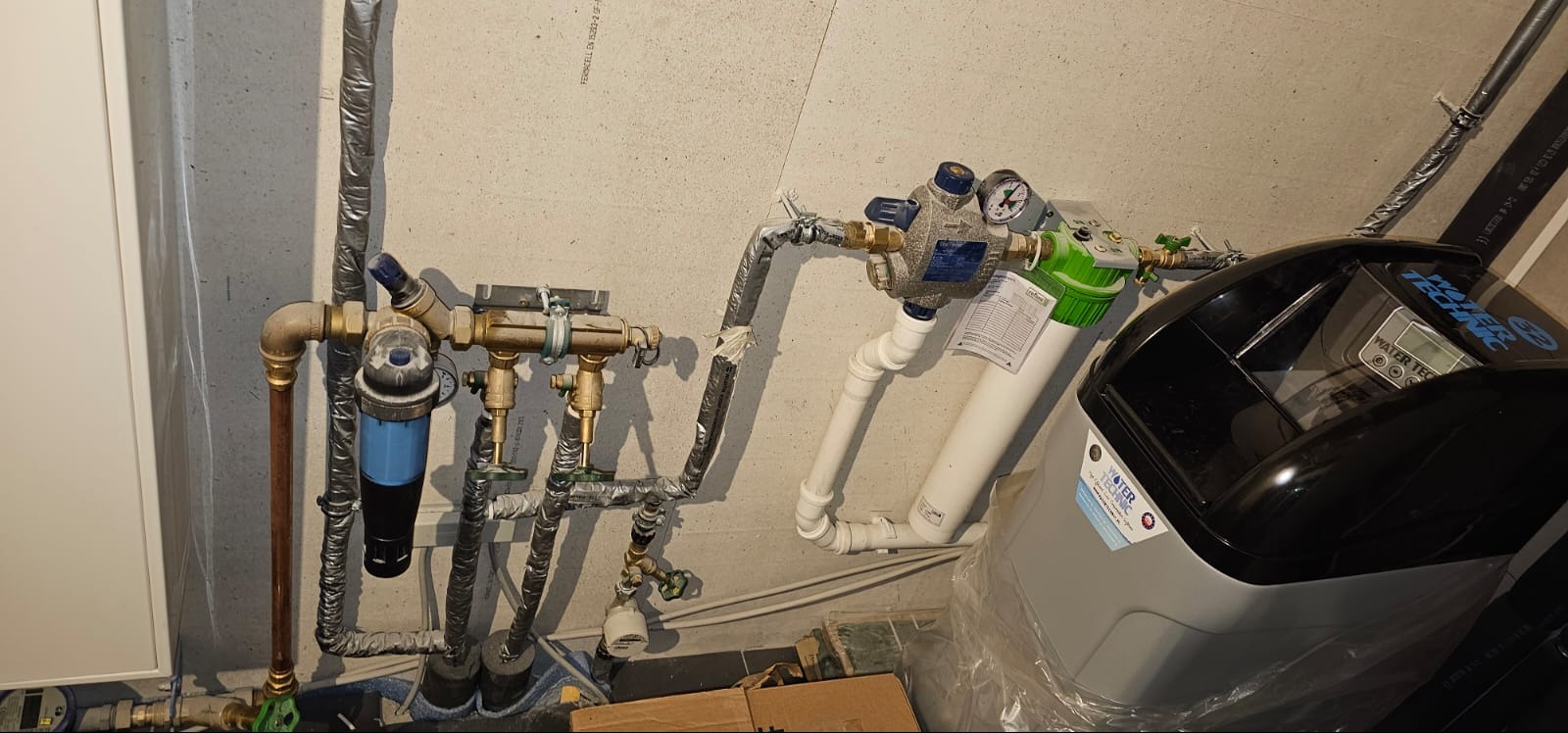 Złożona instalacja wodna z filtrami, zaworami i zmiękczaczem wody Water Technic, rury miedziane i izolowane, szara ściana w tle.