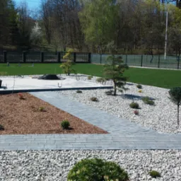 Nowoczesny ogród z trawnikiem, ścieżką z szarych płyt, białymi kamieniami, korą drzewną i młodymi drzewkami, całość otoczona czarnym ogrodzeniem.