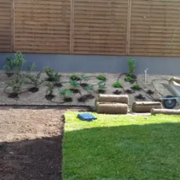 Fragment ogrodu z fragmentem ułożonej trawy z rolki, przygotowanym podłożem i roślinami na zboczu z systemem nawadniania, taczka z rolkami trawy, drewniany płot w tle.