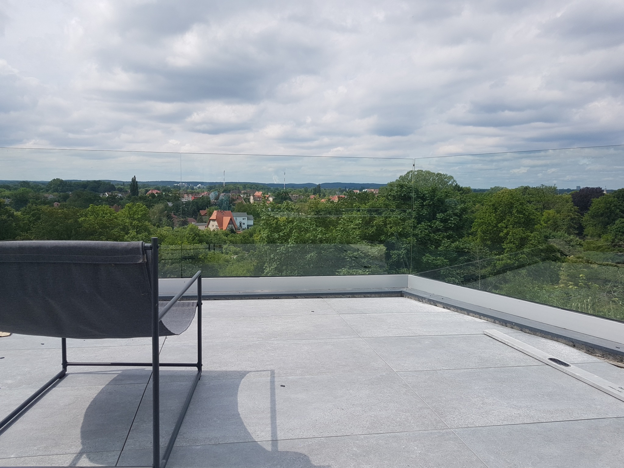 Szare płytki na tarasie z szklaną balustradą i widokiem na zieleń i zabudowania w oddali, z krzesłem w lewym dolnym rogu.