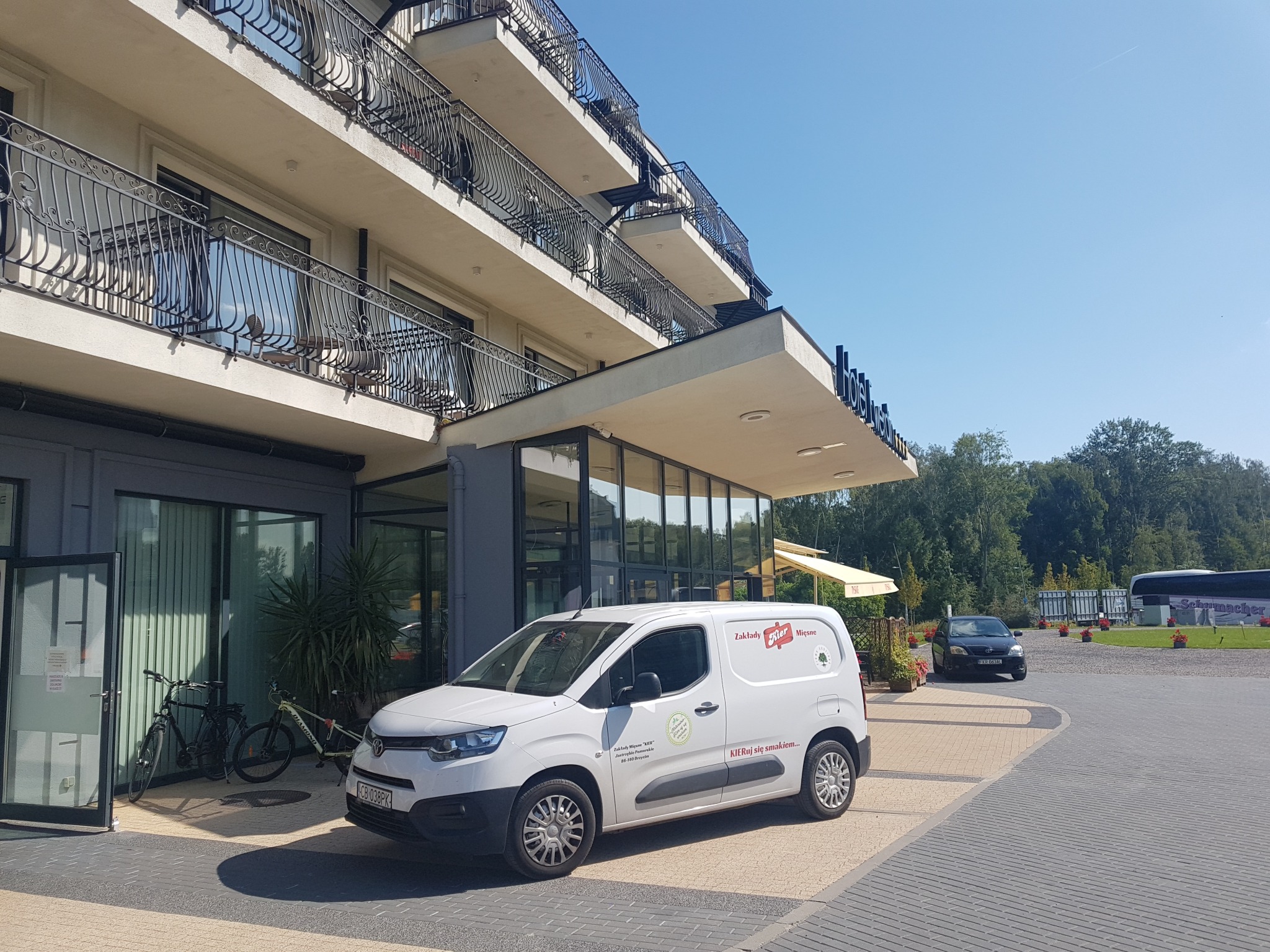 Biały samochód dostawczy z logo firmy Zakłady Mięsne KIER zaparkowany przed budynkiem hotelu z balkonami i przeszklonym wejściem, rowery opierają się o ścianę obok wejścia.