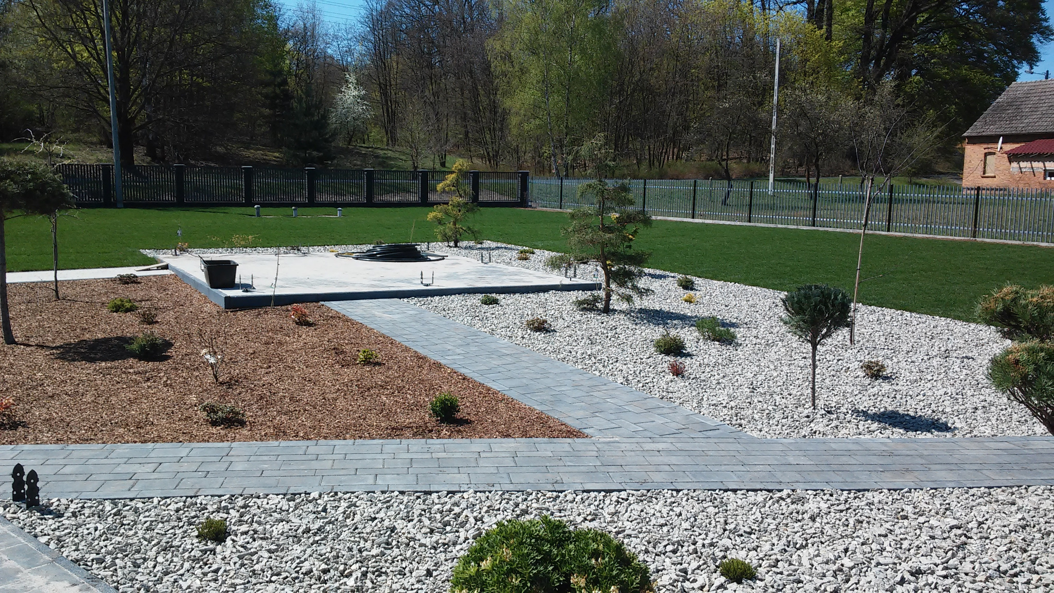 Nowoczesny ogród z trawnikiem, ścieżką z szarych płyt, białymi kamieniami, korą drzewną i młodymi drzewkami, całość otoczona czarnym ogrodzeniem.