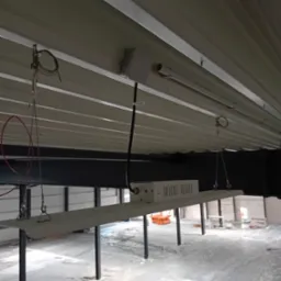 Wnętrze hali w trakcie instalacji oświetlenia: widoczne lampy LED zawieszone na stalowych linkach, kabel zasilający oraz zabezpieczona folią podłoga.