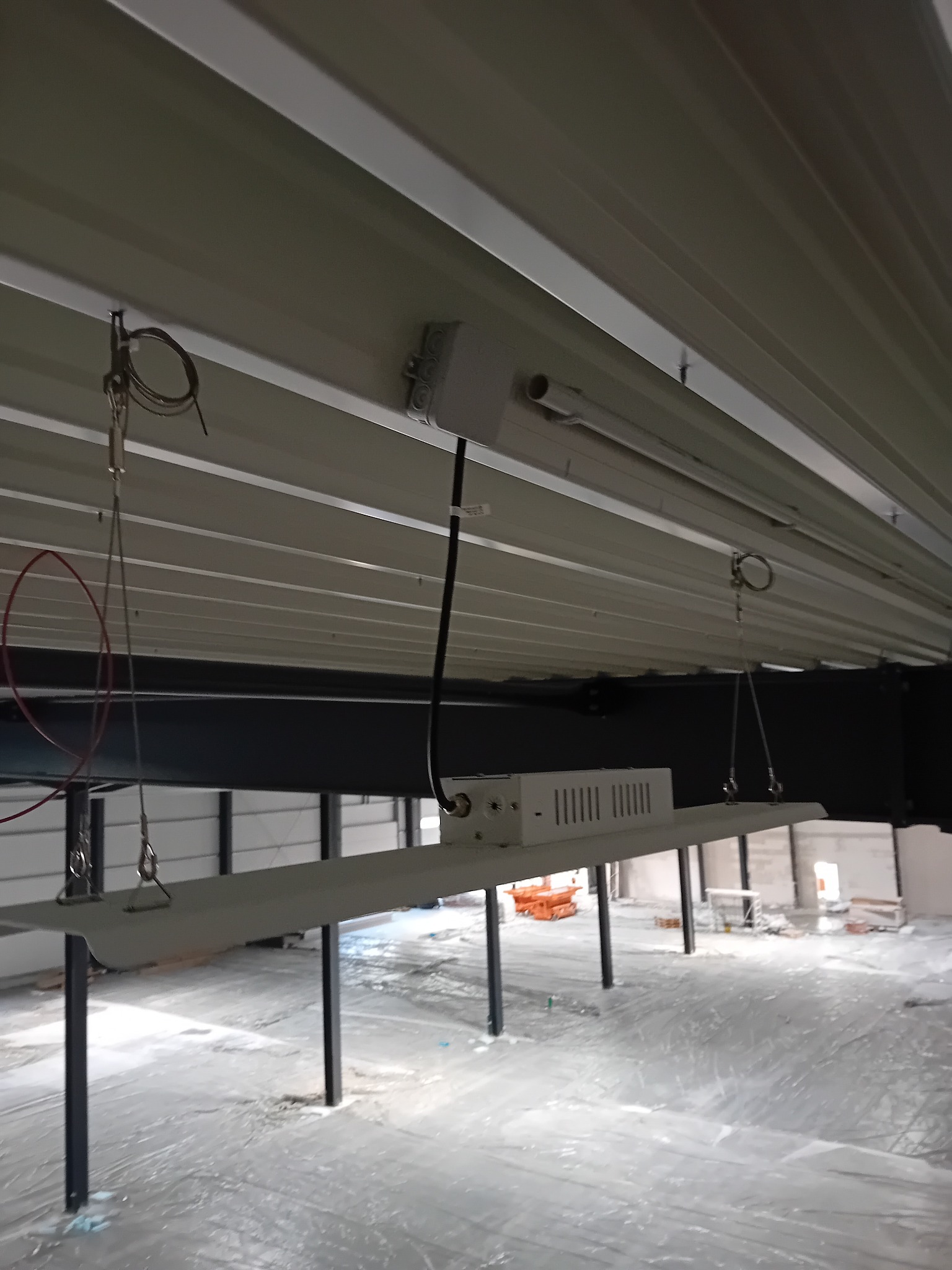 Wnętrze hali w trakcie instalacji oświetlenia: widoczne lampy LED zawieszone na stalowych linkach, kabel zasilający oraz zabezpieczona folią podłoga.