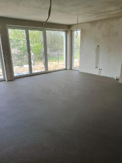 Świeżo wylana, gładka, szara posadzka w surowym wnętrzu z dużymi oknami wychodzącymi na zieleń; widoczne wystające kable z sufitu.
