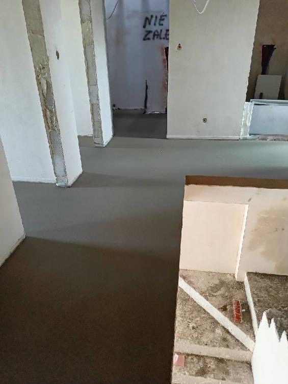 Świeżo wylana, gładka, szara podłoga w surowym stanie, z widocznymi kolumnami i napisem 'NIE ZALE' na ścianie w tle.
