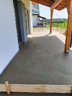 Świeżo wylana, gładka powierzchnia betonowa tarasu przed domem jednorodzinnym, widoczne drewniane szalunki, jasne, słoneczne oświetlenie.