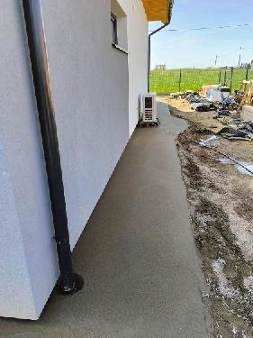 Świeżo wylana, gładka ścieżka z betonu wzdłuż ściany domu z zamontowanym klimatyzatorem, widoczne materiały budowlane w tle.