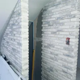 Wnętrze w trakcie prac wykończeniowych: ściana z białej cegły dekoracyjnej z szarymi akcentami, widoczna puszka elektryczna bez osprzętu i poziomica oparta o ścianę. Część ściany w surowym stanie...