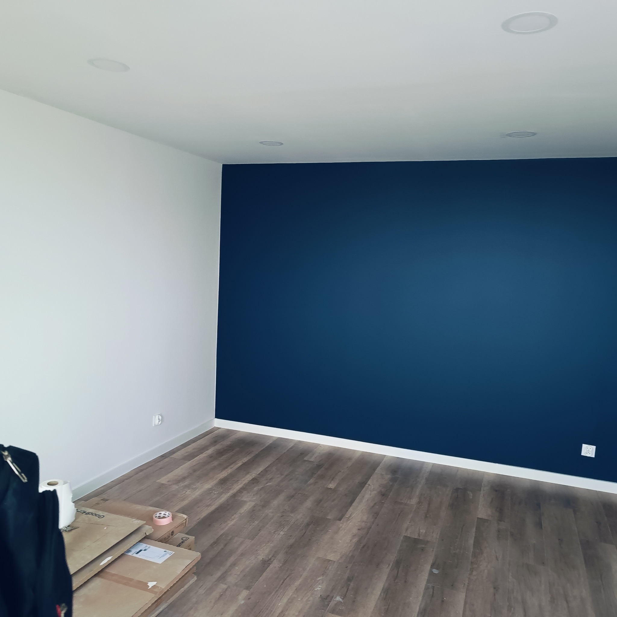 Pomieszczenie po odświeżeniu: ściana pomalowana na granatowo, druga na biało, drewnopodobne panele na podłodze, białe listwy przypodłogowe, kartony i akcesoria malarskie w rogu.