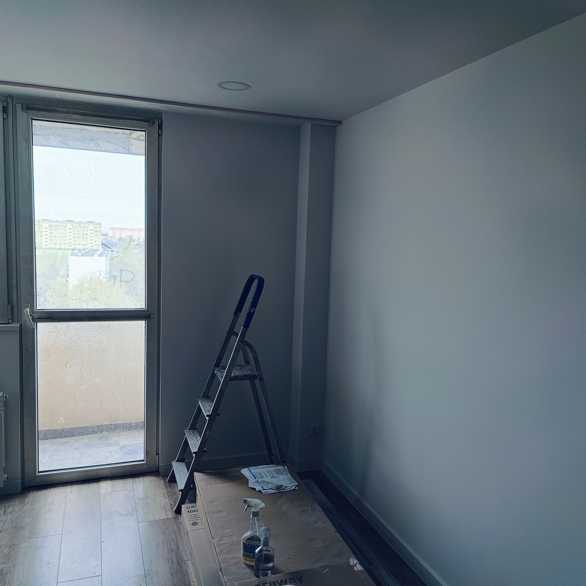 Pokój w trakcie odświeżania, widoczne świeżo pomalowane ściany w jasnym odcieniu, drabina oparta o ścianę, zabezpieczona podłoga i okno balkonowe z widokiem na osiedle.