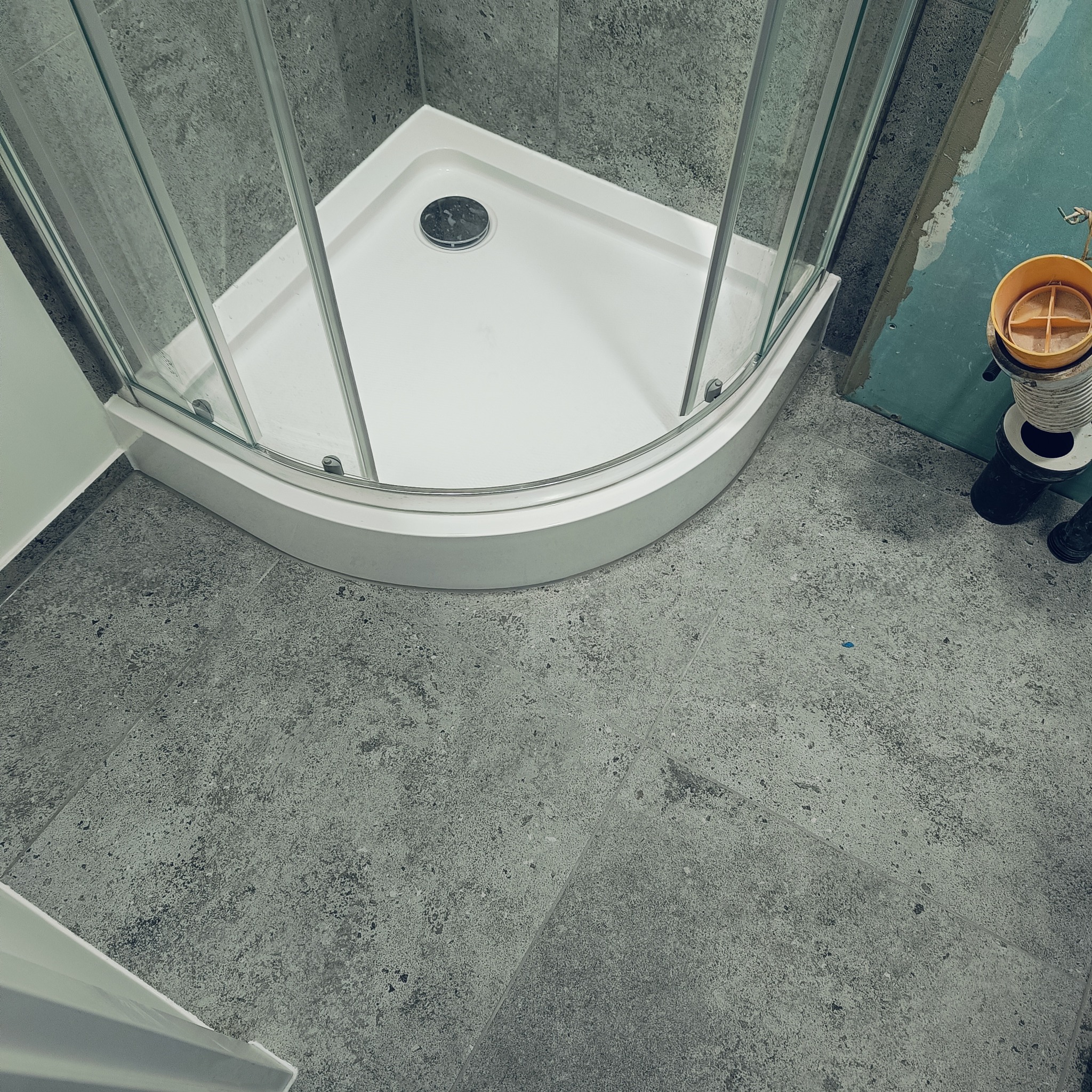 Narożna kabina prysznicowa z białym brodzikiem i szklanymi ściankami, zamontowana na szarej, teksturowanej podłodze z płytek. Widoczny fragment ściany przygotowanej pod wykończenie oraz kubeł...