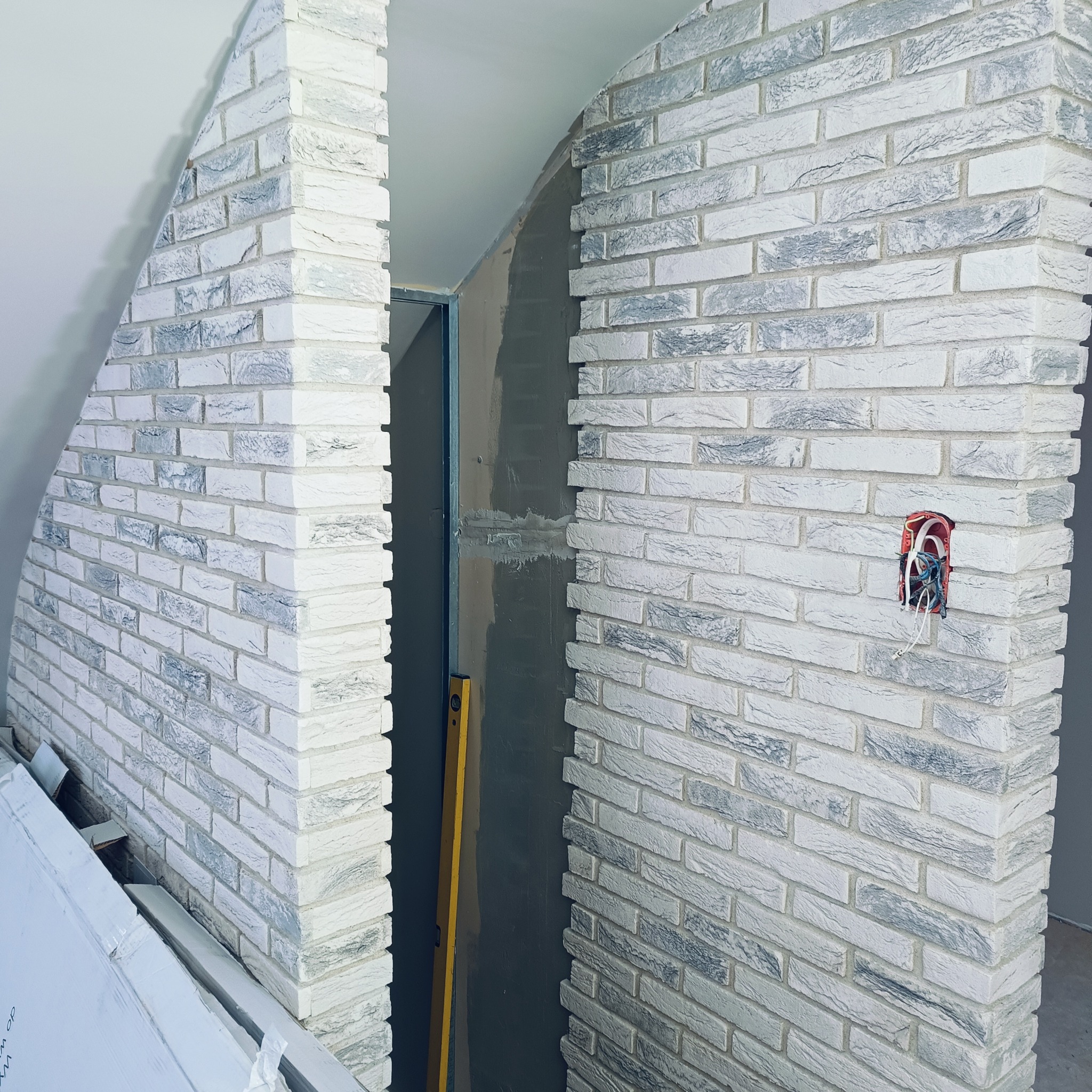 Wnętrze w trakcie prac wykończeniowych: ściana z białej cegły dekoracyjnej z szarymi akcentami, widoczna puszka elektryczna bez osprzętu i poziomica oparta o ścianę. Część ściany w surowym stanie...